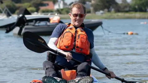 Amy Tallula Will Behenna sat on a paddleboard, paddling on the water with sunglasses on and an orange life jacket