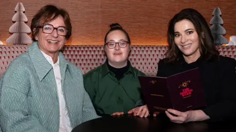 Three women sit at a pub table. They are all smiling. One is holding a menu. 