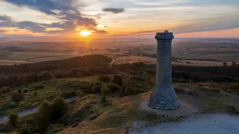 Stone monument on top of heathland with fields stretching into the distance - the sun it just setting.