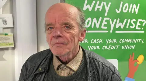 Brian Lindsay standing in front of a green sign which can be partially seen. It says "Why join Moneywise". Lindsay has grey hair and is wearing a black jacket and a yellow checked shirt.