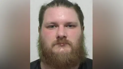 Mugshot of Adam Leddra. He has a large round face with a thick straggly ginger beard and short dark hair pushed backwards.