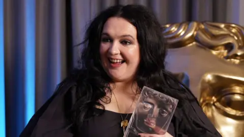 Ashleigh Storrie smiles while holding her Bafta Scotland award. She is wearing a black dress with long puffy sleeves and is standing in front of grey curtains and a large gold Bafta head