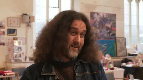 Chair of The Strand Community Trust, Barrie Cooper. He has shoulder length, dark hair which is thick and frizzy, he has a full beard with grey on the chin and dark eyebrows. He's wearing a dark blue denim jacket. Behind him are paintings, cards and tables of craft items.