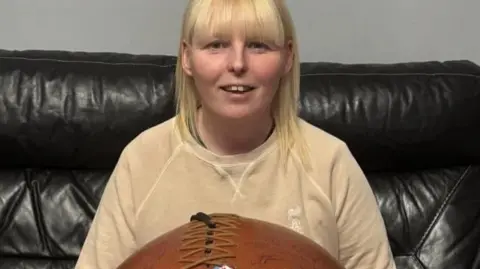 A woman with shoulder-length blonde hair sits on a black sofa, wearing a beige top. She holds a brown ball on her lap with stitches along the top holding the leather together.