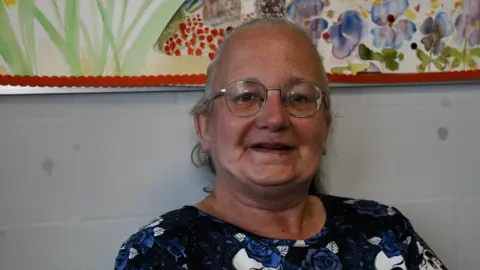 John Fairhall/BBC A head and shoulders image of a woman wearing glasses and a blue, black and white floral top. She is looking into the camera while smiling. 