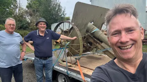 Great War Huts Three men pose with a rusty World War One howitzer gun on a trailer. The gun is in the middle of them and has been strapped to the trailer. 