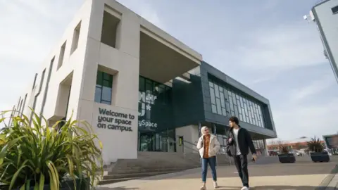 A building part of the city campus of the University of Sunderland, with two young people, a man and a woman, walking away from the building. The building has a main white block, with a black-clad windowed section to one side. Steps lead up to a glass-fronted entrance.