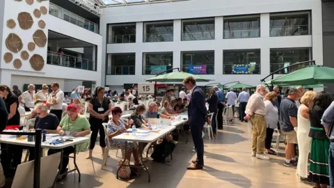 A crowd of people in an atrium, gathering around people sitting at trestle tables counting vote papers