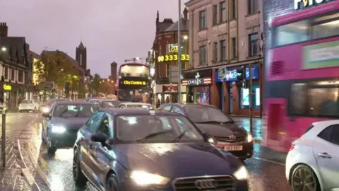 Queues of cars and buses are pictured in Belfast city centre. It is evening time and the lights of the cars as well as shop signs along the streets are lit up.