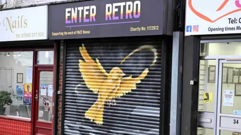 Karbon Homes A gold bird is painted spreading its wings on the balck shutters of Enter Retro. There is a nail salon with a red shop front next to the premises.