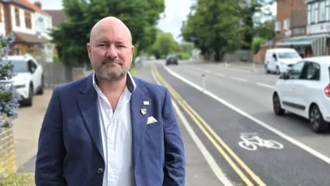 Paul Moseley/BBC A man in a blue blazer stands on a suburban roadside, facing the camera with hands in pockets. To the left, a flowering shrub borders a low brick wall and houses. To the right, a street with a bike lane, passing cars, and shops stretches into the distance beneath leafy trees and overcast light.