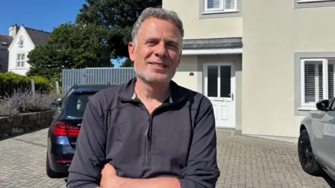 Nick, who has short grey hair and a beard, wearing a blue zip top. He has his arms crossed in front of him and is smiling at the camera, with a cream coloured house behind him and two cars on the driveway.