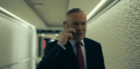 5 Broadcasting Limited / Paramount / Wonderhood Studios/ PA Clunes in a suit with a light blue shirt and red tie. Holding a phone to his right ear