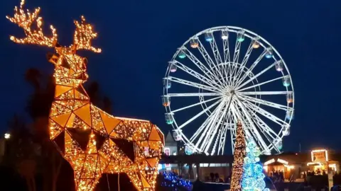 An illuminated reindeer made up of triangle shapes and covered in warm gold lights. In the background is a large white ferris wheel, also lit up against the night sky.