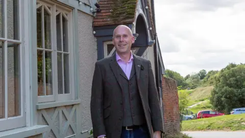 Supplied David Scott standing outside a building and in front of a green, hilly backdrop. He is wearing a dark coloured suit jacket over a waistcoat and a pink shirt. He is smiling and looking into the camera. 