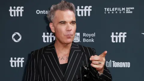 Reuters Singer Robbie Williams wearing a black jacket with slim, white stripes, points to the right of the frame while speaking to someone off camera.