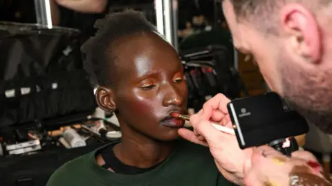 Getty Images Arop Akol, wearing a dark green sweater, gets colourful make-up done by a make-up artist backstage at the Annie's Ibiza Collection show during London Fashion Week - February 2025.