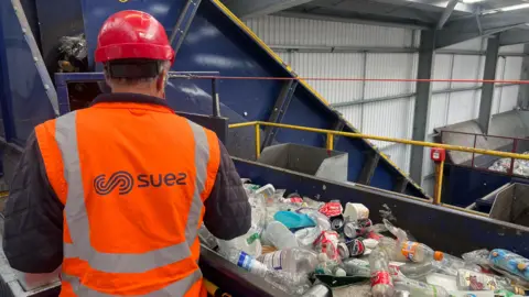 BBC Recycling centre worker with his back to the camera with a high-viz jacket on saying 'Suez'