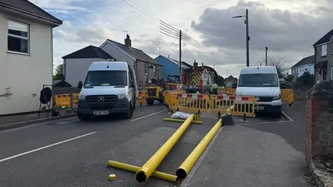 BBC Two yellow poles laid on the tarmac in front of a construction site with two white vans parked either side