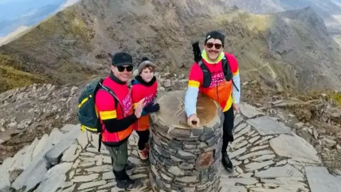 The Bavington family including 8-year-old Harry stand by the summit cairn marker on the top of Snowdon (Yr Wyddfa) they are wearing bright pink and orange shirts with Breast Cancer Now written on them.