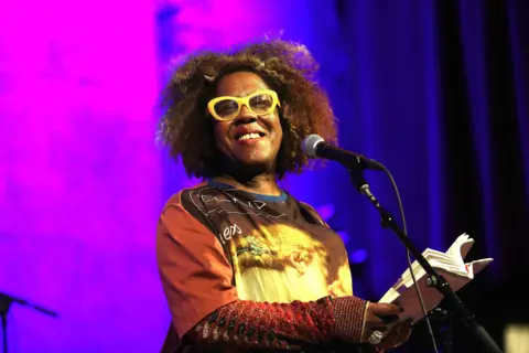 Getty Images Errollyn Warren performs in front of a microphone with a book in hand. He wears yellow glasses and a yellow and brown T-shirt over a long-sleeved reddish top. The background is blue and pink.