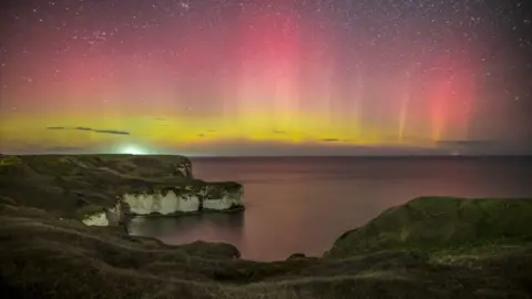A rugged coastal cliff overlooks calm water beneath a sky illuminated by vivid bands of red, pink and yellow light from a striking aurora display. Stars scatter across the night sky above the glowing horizon, creating a dramatic contrast with the dark landscape below.