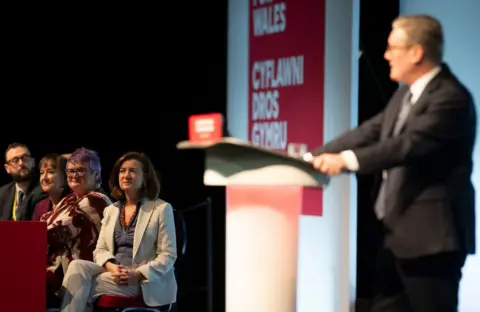 Getty Images A man with grey hair, glasses, a navy suit, blue shirt and a red tie - Sir Keir Starmer - stands at a podium while giving a speech. In the background sits a woman with medium length brown hair wearing a white suit - Eluned Morgan - alongside two women and a man.