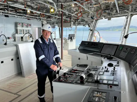 David Tydeman, a middle aged man dressed in blue overalls and a white helmet stands on the bridge of Glen Sannox, holding the wheel that steers the ship. The control equipment for the ship can be seen in front of him and the Clyde is visible through the bridge windows.