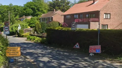 A road junction with two warning signs - one, painted yellow, says 'no access to Whitby,' the other, painted red, says 'road closed one mile ahead.' There is also the classic 'roadworks' sign, showing a silhouetted figure with a spade.