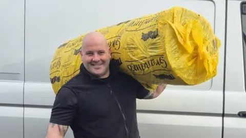 Supplied Stuart holds a wrapped up roll of carpet on his shoulder. He is wearing a black top and grey shorts. He has black socks and black trainers. He is smiling and holding a carpet fitting tool. There is a silver van behind him.