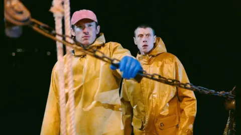 The image shows two men are dressed in bright yellow oilskin jackets. It is pitch black behind them. The man in the foreground is holding a chain and ship's ropes hang vertically in front of him. The second man has his mouth open in shock.