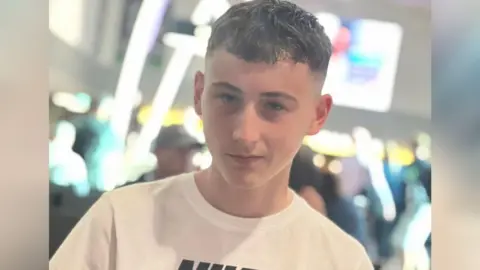 Gemma Kirk Jack, a boy with short black hair, wearing a white t-shirt. He is looking at the camera. There are lights and people in the background, which is out of focus.