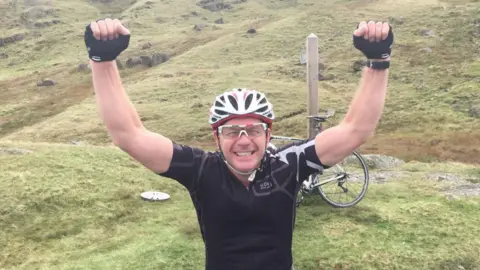 A man in a black cycling top and white cycling helmet with both arms held up in the air and a bicycle behind him on the grass