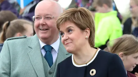 PA Media Peter Murrell and Nicola Sturgeon earlier  astatine  an lawsuit   their separation. Murrell has a bald caput  and is wearing thin-framed glasses, a greenish  overgarment   and waistcoat with a achromatic  garment  and bluish  tie. Sturgeon is smiling and is wearing a bluish  dress. There are children successful  the background