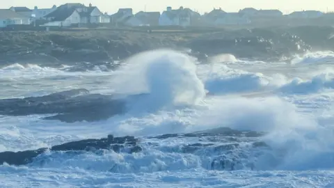 Ann 97 Waves crashing on Trearddur Bay, off the coast of Anglesey. Houses on the coastline can be seen in the background.