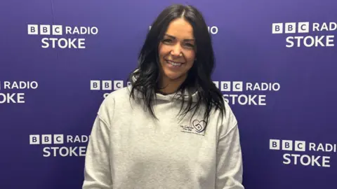 BBC A woman with long black hair and a light grey hoodie smiles as she stands in front of a purple background with the BBC Radio Stoke logo on it.