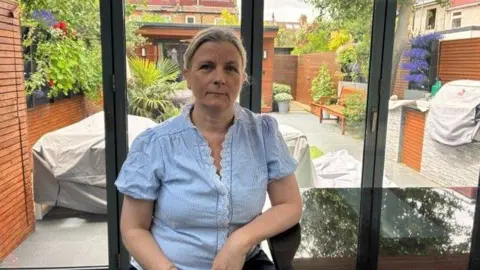 Sabine with fair hair tied back, wearing a light blue striped top, sitting in front of open glass doors that look onto a garden.