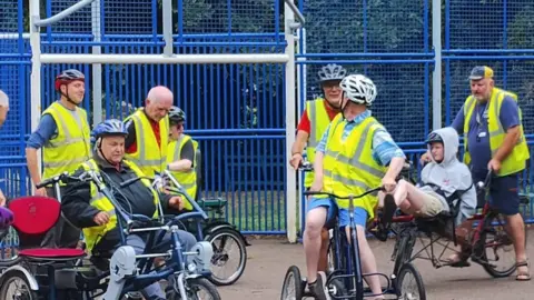 Contributed Cyclists wearing hi-vis jackets stand around inside a playground before they are about to embark on a cycle ride. The talk among each other.