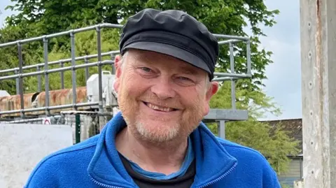 Tom Jackson/BBC David Goode is standing in front of the closed lock at Baits Bite. Behind him is a large red 'lock closed' sign. David is wearing a blue fleece, black cap and black top. He has grey and ginger facial hair.