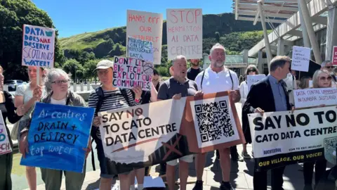 A group of people stand outdoors in bright sunshine holding protest signs opposing a data centre development. The group is gathered near a modern building with a metal canopy, with green hills and a clear blue sky in the background.
