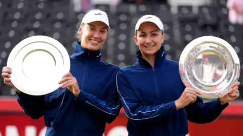PA Media Tereza Mihalikova (left) and Olivia Nicholls (right) standing next to each other, each holding their Queen's women's doubles trophy. They are both looking directly at the camera and are smiling.