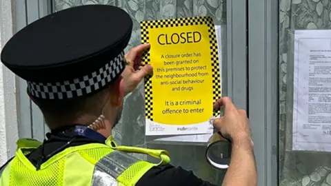 A male police officer is attaching a yellow "closed" notice to a front window of a house. The window has net curtains and another explanatory notice is attached to the other side of the window. The officer is wearing a police hat and a hi-vis yellow jacket with the word "police" across the back of it
