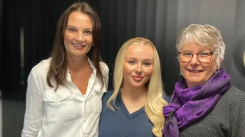 BBC Left to right:  Sam, Bath and Anne. Sam has long brown hair and is wearing a white shirt. Beth has long blonde hair, a nose ring and is wearing a navy V-neck top. Anne has short grey hair and is wearing a grey top and is wearing a purple scarf 