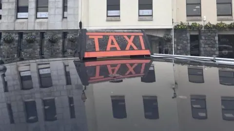 A red taxi sign on top of a taxi which is reflected in the shiny black surface of the vehicle