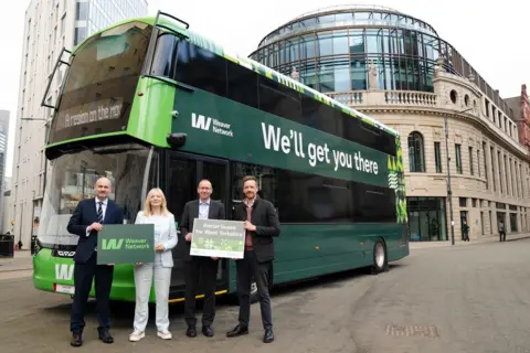 Four people stand in front of a green double-decker bus with buildings behind. The people are holding two banners that read 'Weaver Network' and 'Better buses for West Yorkshire'. The sign on the side of the bus reads 'Weaver Network: We'll get you there'.