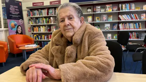 Clare Worden/BBC Edith Reeves is wearing a fluffy brown coat. She is sitting in the library with shelves of books behind her. 