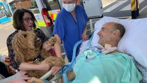East Kent Hospitals University Trust Martha's paw shaking Nigel Avery's hand while he is on a hospital bed