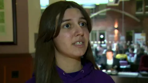 Emma, with long brown hair and wearing a purple hoody, looks to the side of the camera