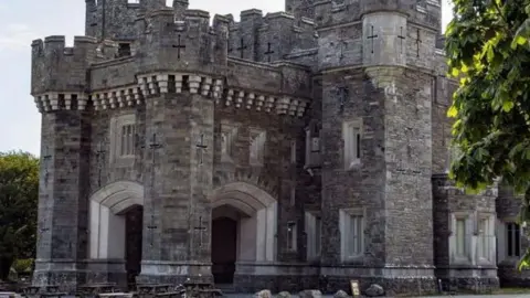 Hugh Mothersole Wray Castle which is a large, grey stone gothic-looking castle with turrets. There are brown picnic benches in front of the arched entrance. Trees are on either side of the building.