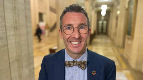 Andrew Muir. He has shirt grey-ish hair and blue eyes, wearing glasses, a blue blazer, blue check shirt and yellow check bow tie.